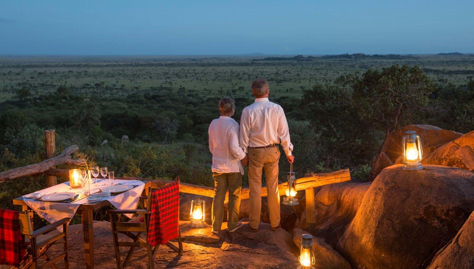 A couple watching the savanna at dusk beside a candlelit dinner table and lanterns