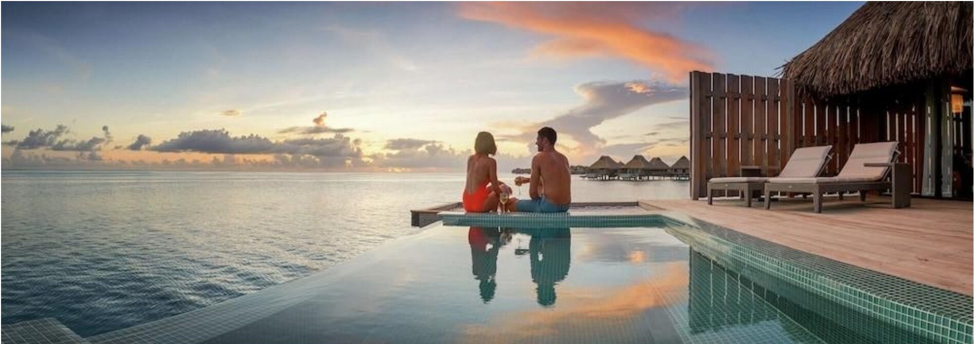 A couple by an infinity pool at sunset overlooking overwater bungalows.