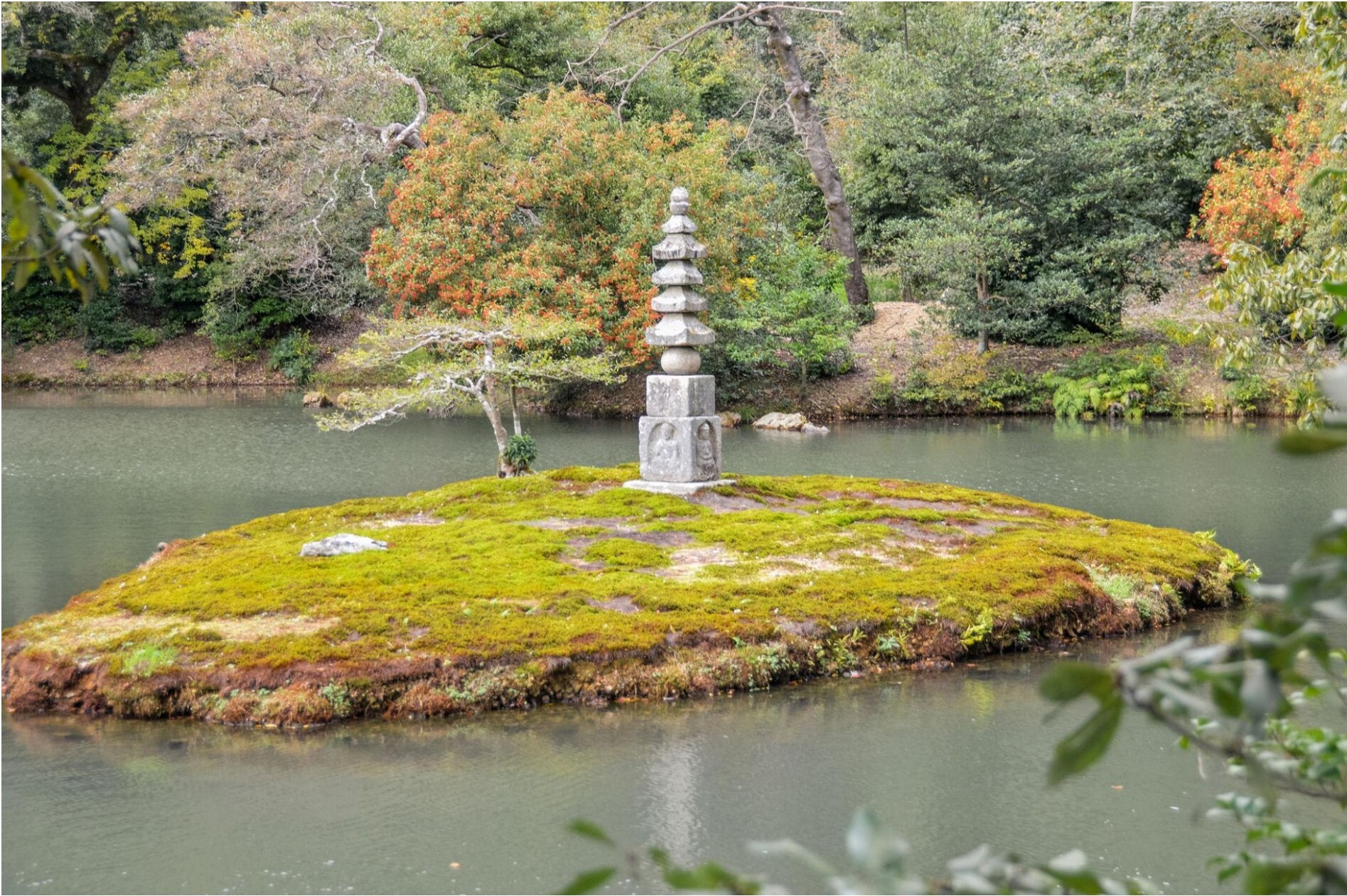 A stone pagoda on a mossy island in a Kyoto temple pond, surrounded by autumn foliage.