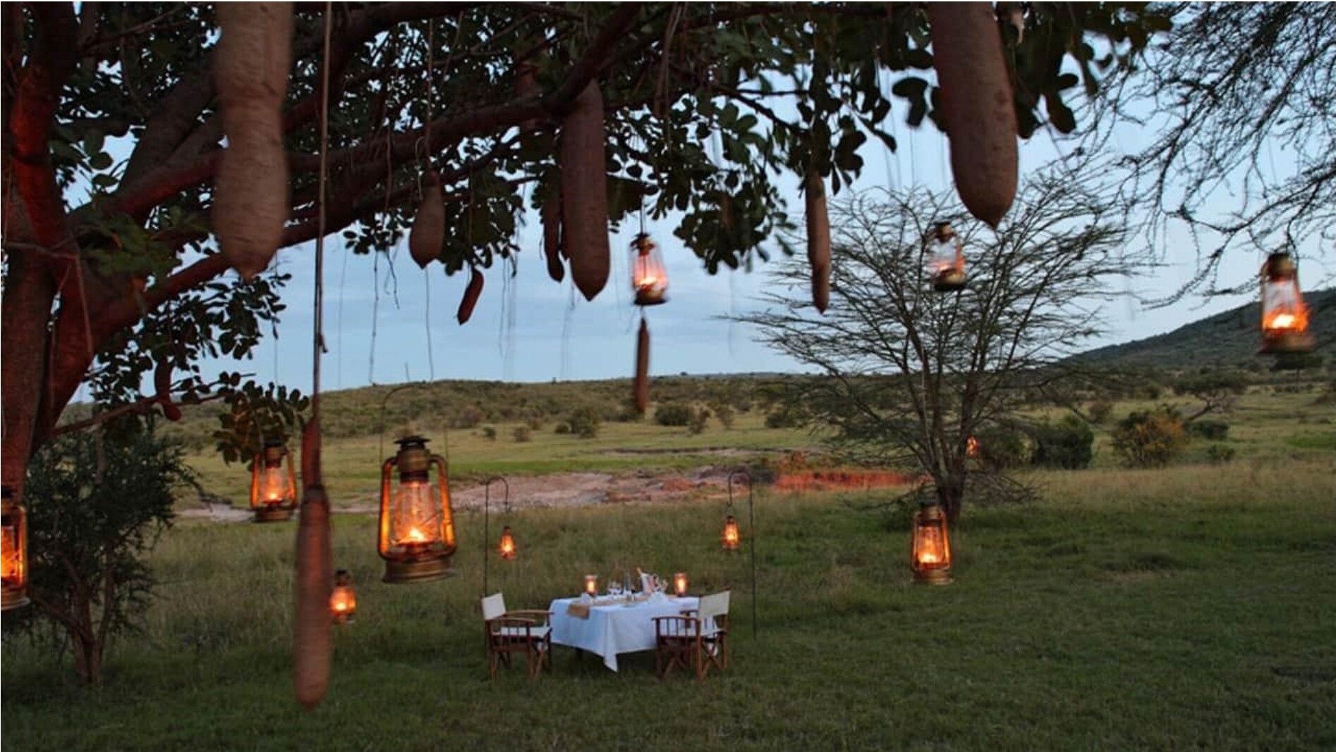 A lantern-lit camp at dusk with a dinner table in the grass