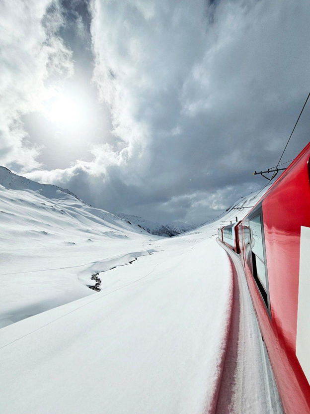 View from a red train winding through a vast snow-covered Alpine valley under dramatic skies.