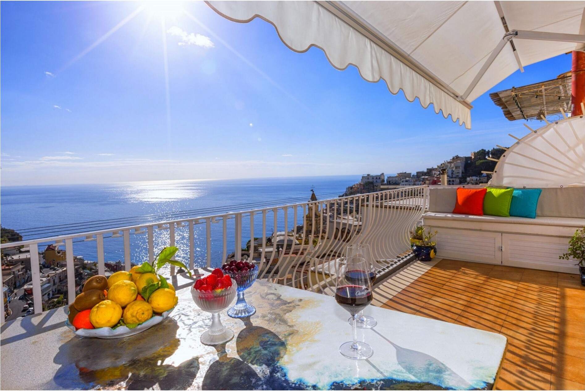 A scenic terrace overlooking the sea with lemons, wine, and fruit on a table.