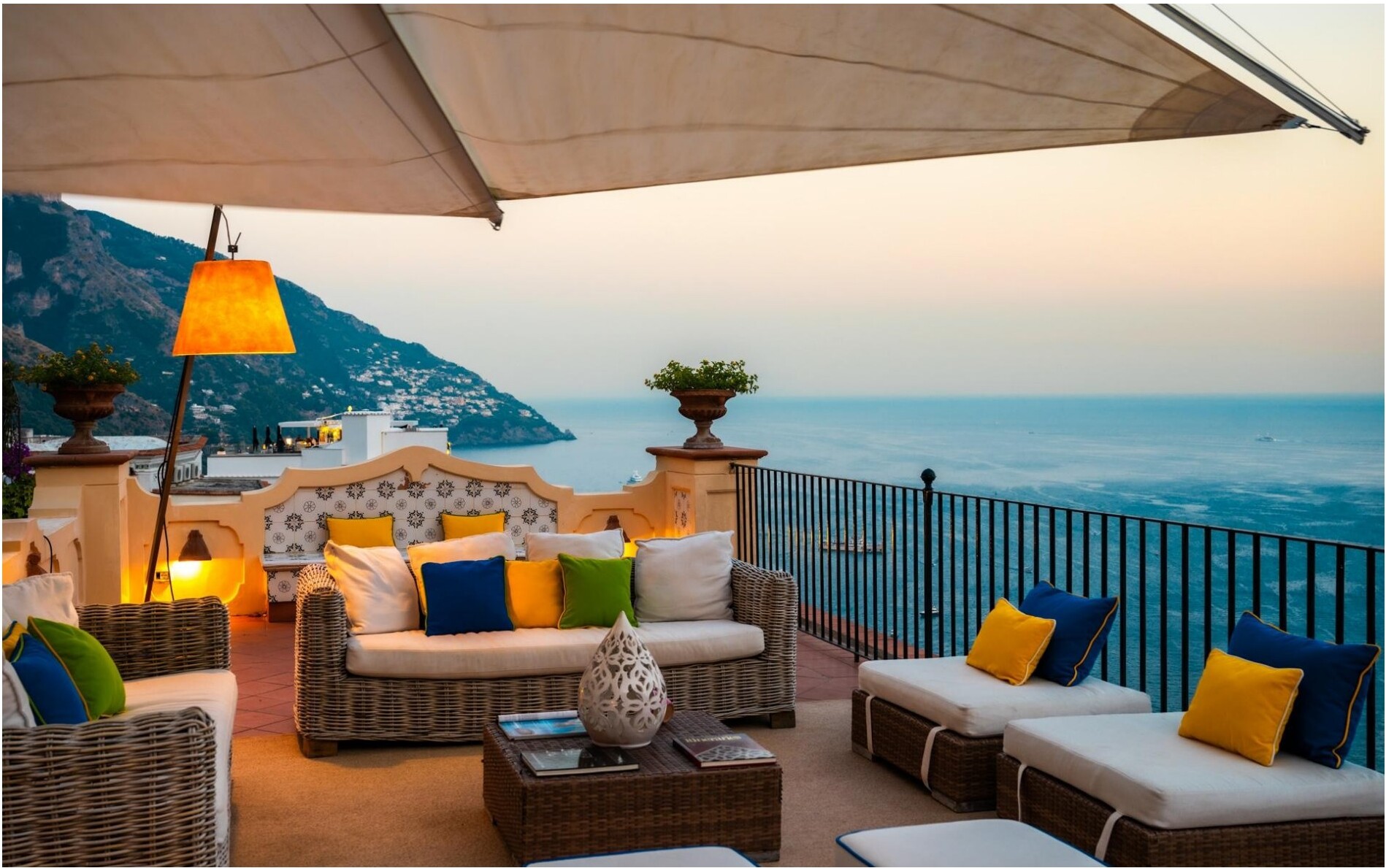 A terrace with wicker furniture, colorful cushions, and a view of the sea at sunset.