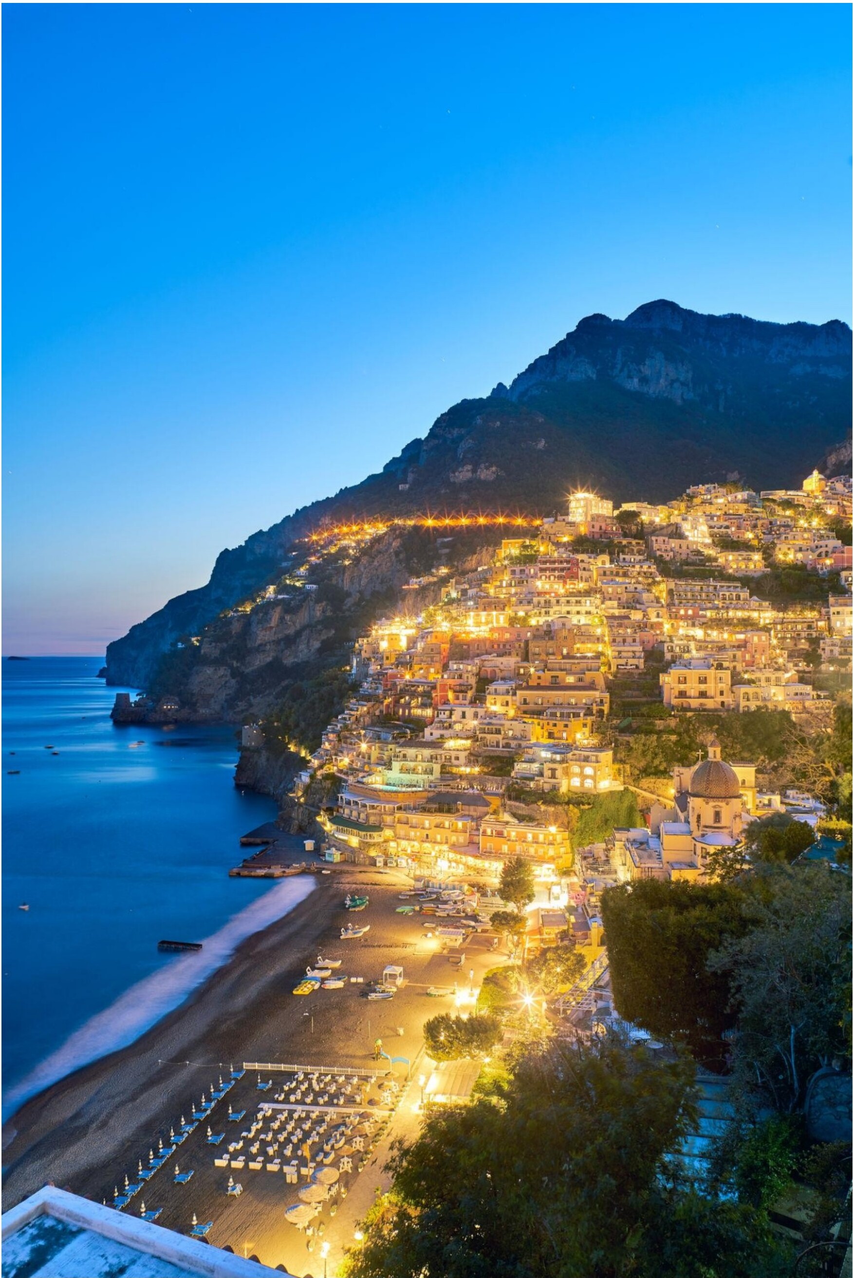 A coastal town at dusk with illuminated buildings cascading down a hillside.