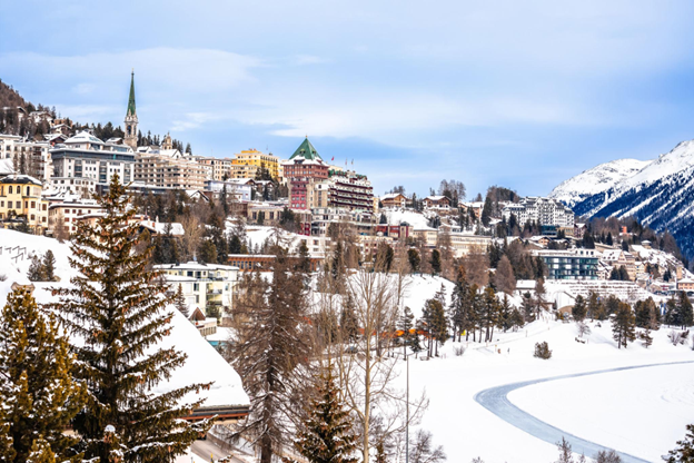 A snow-covered Alpine village with a church spire and mountains in the background.
