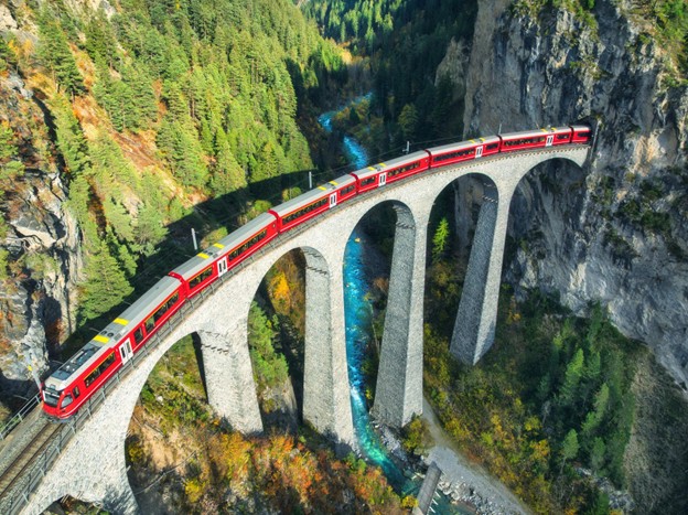 A red train crossing a stone viaduct in a forested mountain landscape.